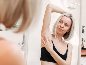 Blonde Frau mittleren Alters mit schwarzem Bustier vor dem Spiegel bei Selbstuntersuchung der Haut unter dem Achselbereich