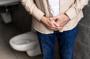 Älterer Mann mit vor dem Bauch zusammengelegten Händen vor Toilette