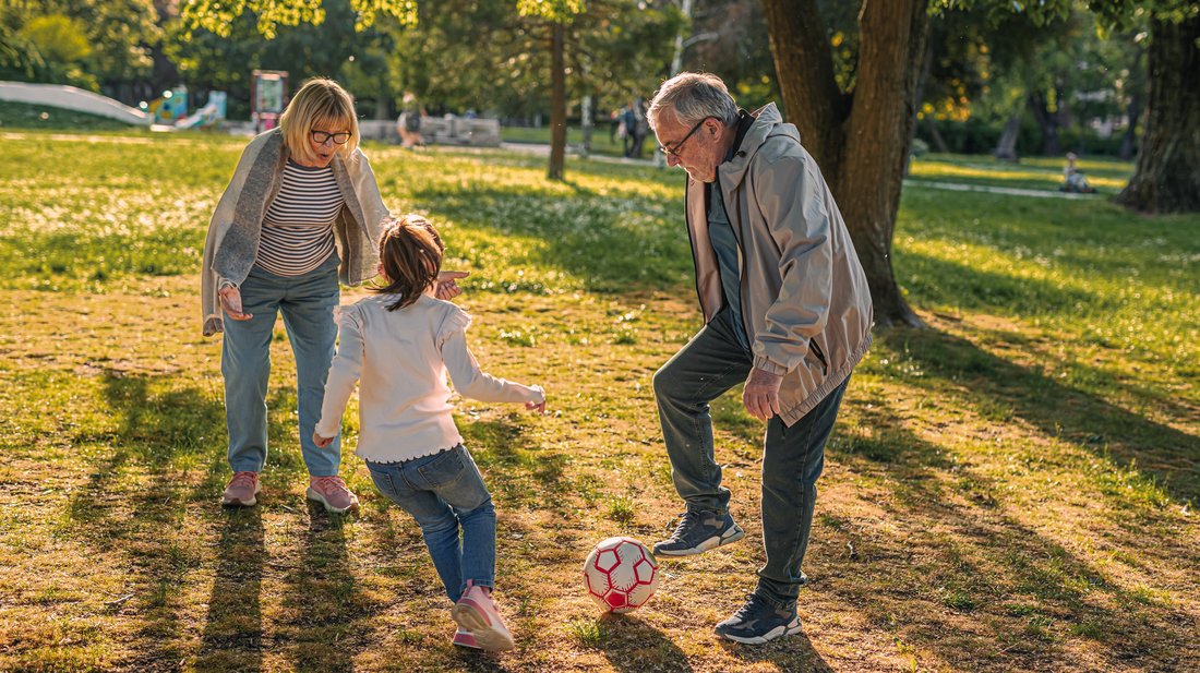 Großeltern spielen mit Enkelin auf einer Wiese mit Bäumen im herbstlichen Sonnenschein