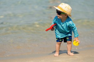 Kind mit Sonnenhut, Sonnenbrille und langärmeliger Oberkörperbekleidung am Strand mit Strandspielzeug
